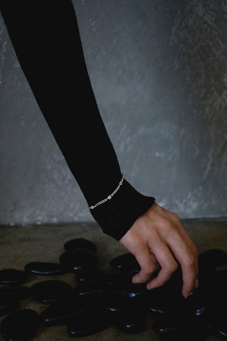 Close up of a woman wearing a black long sleeve top with a sterling silver bracelet layed on top, picking a polished black stone off the floor. 