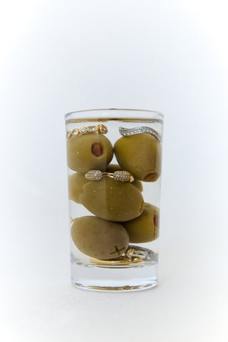 A glass filled with olives and multiple sterling silver and 14k gold vermeil rings with white sapphore accents against a white background. 