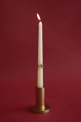 A lit candle in a bronze holder with stacked rings on the candle stick against a red background.
