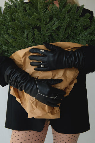 Woman wearing a black blazer, polka dot tights, and black leather gloves, holding a wrapped bundle of christmas tree branches, showing the silver Ryder Ring and sterling silver Reign hand chain.