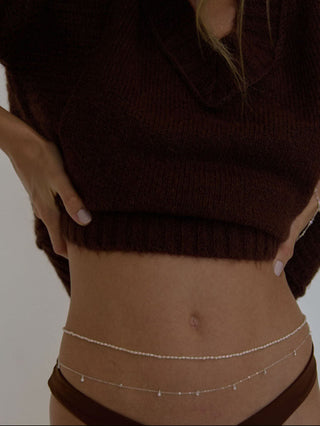 A woman wearing a brown fuzzy sweater and brown bikini bottom with her ams on her hips, showing the double stranded Pearl and sterling silver belly chain. 