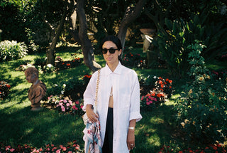 Woman wearing a white button up standing outside amongst trees and flowers