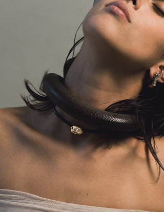 Close-up of a woman's neck wearing a braided leather chocker with gold pendant featuring a white sapphire with a black snake draped over her shoulder against a neutral background