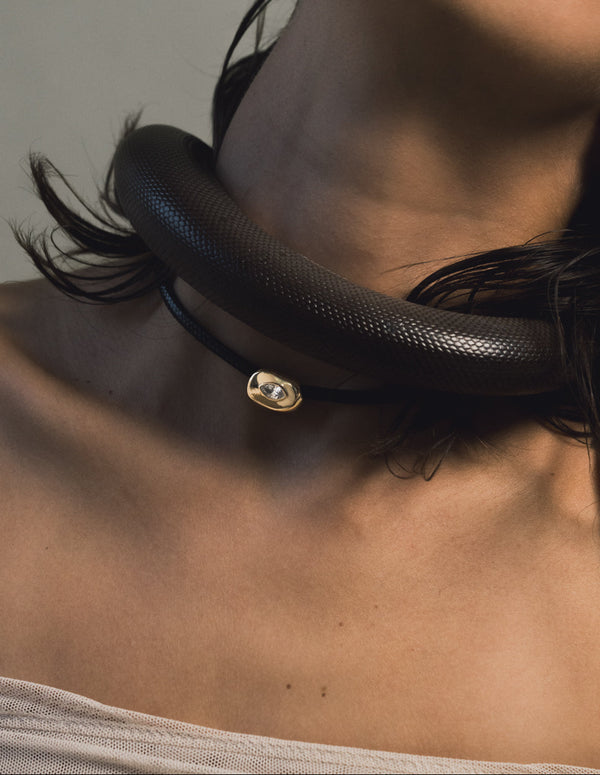 Close-up of a woman's neck wearing a braided leather chocker with gold pendant featuring a white sapphire with a black snake draped over her shoulder against a neutral background
