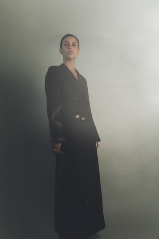 Person wearing a long black coat standing against a plain background with a snake wrapped around her arm and wearing two black leather belts with gold buckle accents. 