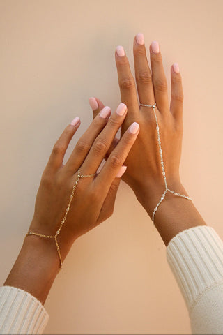 A pair of delicate gold cz hand chains worn on crossed hands, set against a solid beige background.