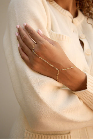 A close-up of a person wearing a delicate gold chain hand bracelet over a cream-colored knit cardigan.
