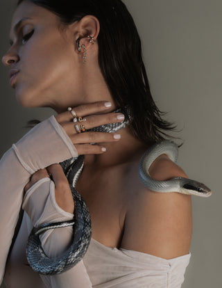 Woman wearing gold and silver rings and earrings with a snake draped over her shoulder against a neutral background