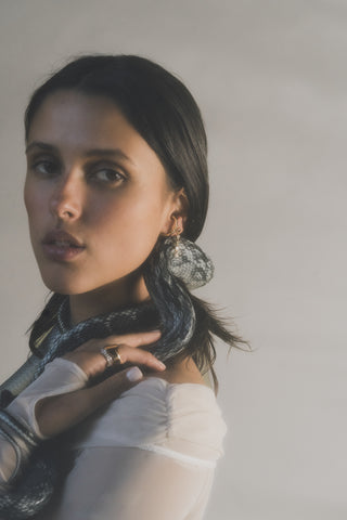 Woman wearing a sheer top and gold and silver rings and earrings with a snake wrapped around her neck and torso. 