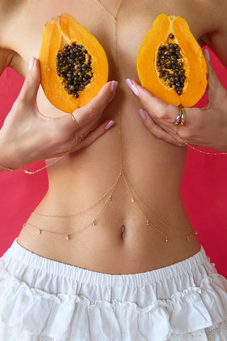 Person wearing godl body chain dn hand chain holding two halves of a papaya in front of their chest against a pink background