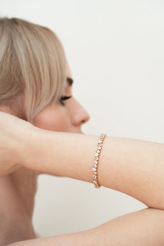 Blonde woman wearing a gold tennis bracelet with lab grown white sapphires on a plain background
