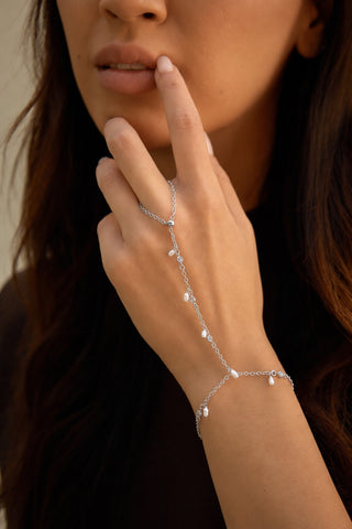 A close-up of a woman wearing a delicate silver hand chain bracelet with dangling pearls, with her hand resting near her face.
