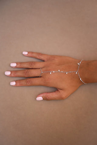 A delicate silver chain hand bracelet with small pearl beads, shown on a model's hand against a solid taupe background.