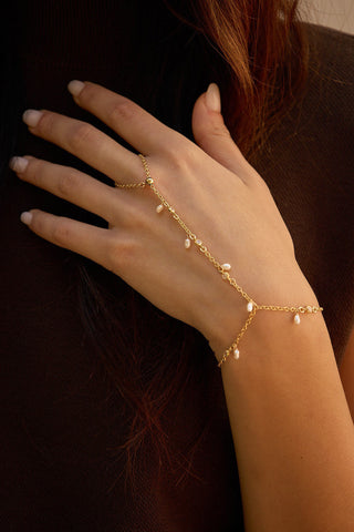 A gold chain hand bracelet with small pearlS worn on a woman's hand and wrist, shown against a dark brown background.