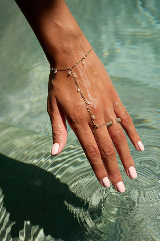 A delicate gold  hand chain with pearls worn on a woman's hand, which is resting in clear, sunlit pool water.