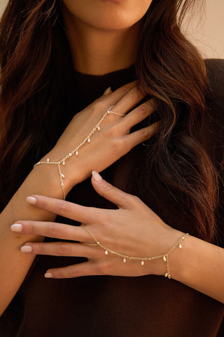 A close-up of a woman wearing delicate gold chain hand chain with small dangling bead accents, styled over a dark brown top.