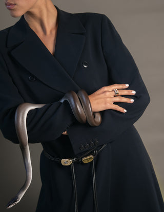 Person wearing silver and gold rings and black leather corded belts with gold buckle accents in a black coat holding a snake with a neutral background