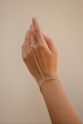 A delicate mixed metal chain hand bracelet worn on a hand, shown against a solid beige background.