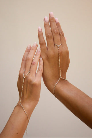 A pair of delicate gold-tone chain hand chains worn on two hands against a solid beige background.