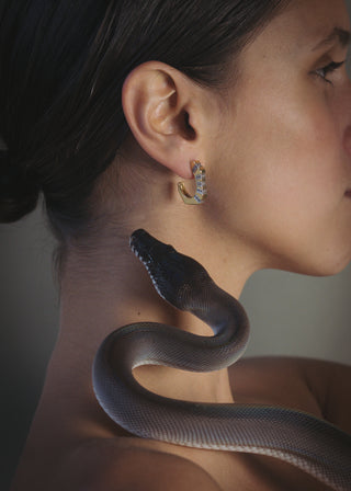 Woman wearing gold hoop earrings with a snake draped over her shoulder against a neutral background