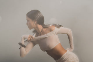 Woman wearing a black leather corded necklace with a gold pendant, gold earrings and rings in a sheer top with a snake across her shoulders against a blurred background. 