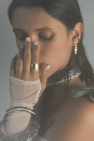 Woman wearing gold rings and earrings with a snake around her shoulders, against a neutral background.