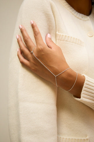 A delicate silver chain hand worn by a model in a cream-colored knit sweater. The minimalist design features a thin chain connecting the ring finger to the wrist, with a small central connector.