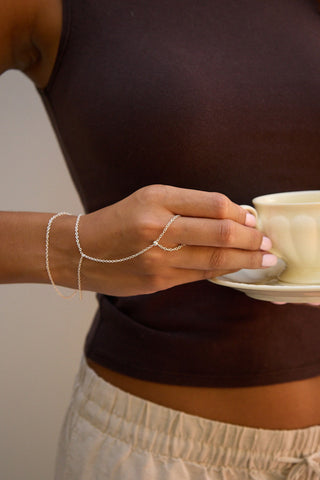 A close-up of a person wearing a delicate silver chain bracelet that wraps around their wrist and hand, while holding a cream-colored ceramic teacup and saucer.