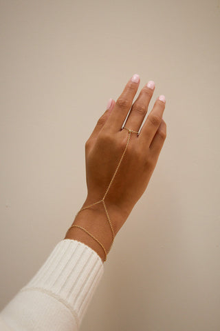 A delicate gold chain hand bracelet worn on a hand with light pink manicured nails, set against a plain beige background.