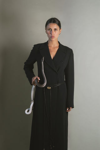Woman in a black coat holding a snake against a gray background wearing black leather corded belts with gold buckle accents.