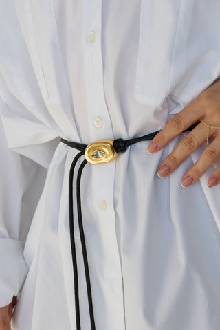 Close-up of a black braided belt with a gold pendant cinching a white button-down shirt at the waist