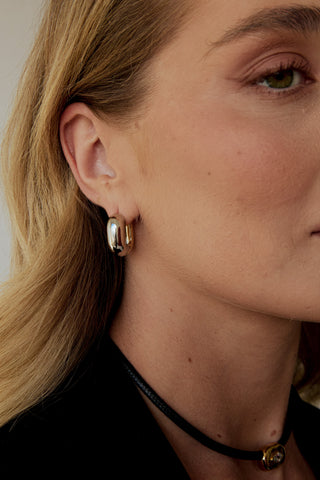 Close-up of a person wearing silver and gold hoop earrings and black corded necklace with a neutral background