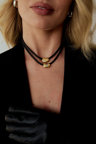 A close-up of a model wearing a black multi-strand cord choker necklace with two polished gold oval charms, styled with a black blazer and a black satin glove.