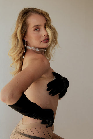 Woman wearing a pearl choker, pearl drop earrings, black gloves and a patterned tights against a plain background