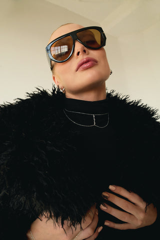 Person wearing large sunglasses, silver choker necklace, and a black feathered garment against a neutral background