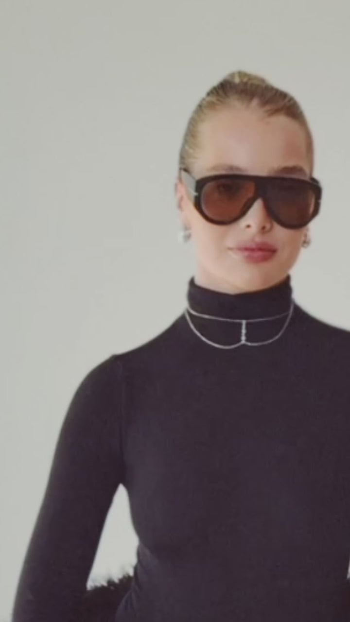 Woman wearing black turtleneck, oversized sunglasses, and silver choker walking, smiling, and picking up presents form the floor