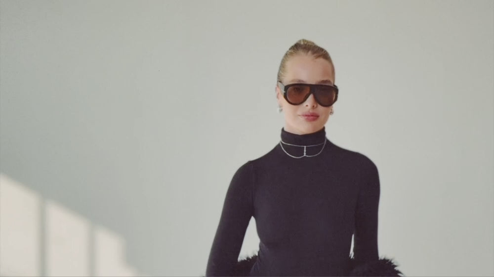 Woman wearing black turtleneck, oversized sunglasses, and silver choker walking, smiling, and picking up presents form the floor